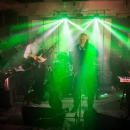 Zespół muzyczny na scenie, oświetlony zielonymi reflektorami, w trakcie występu; wokalista w ciemnej kurtce, basista w białej koszuli, perkusja w tle.