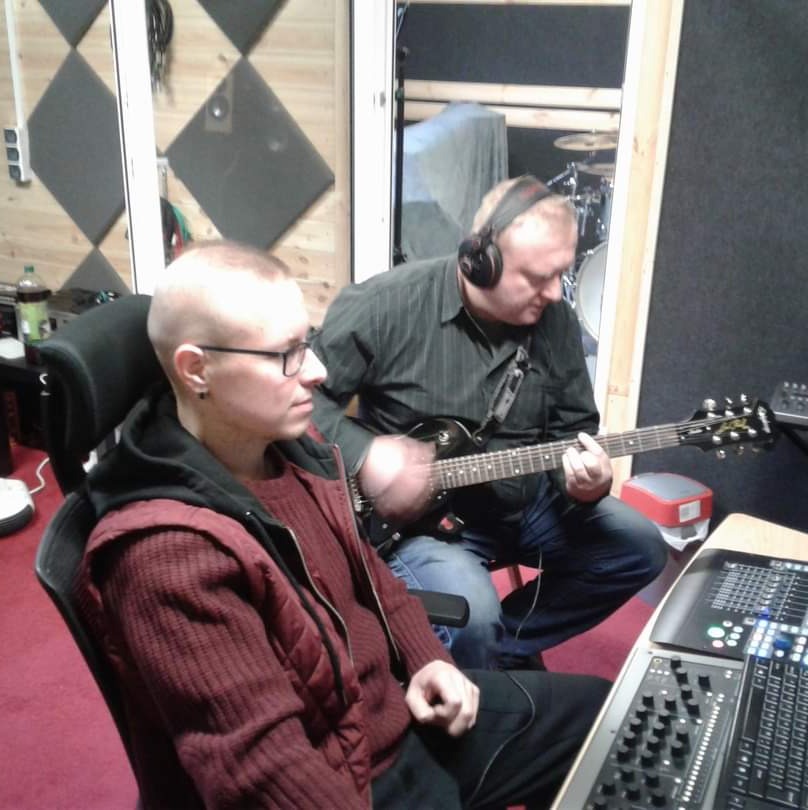 Dwóch mężczyzn w studiu nagraniowym: jeden siedzi przy mikserze, drugi gra na gitarze elektrycznej z słuchawkami na uszach, w tle panele akustyczne.