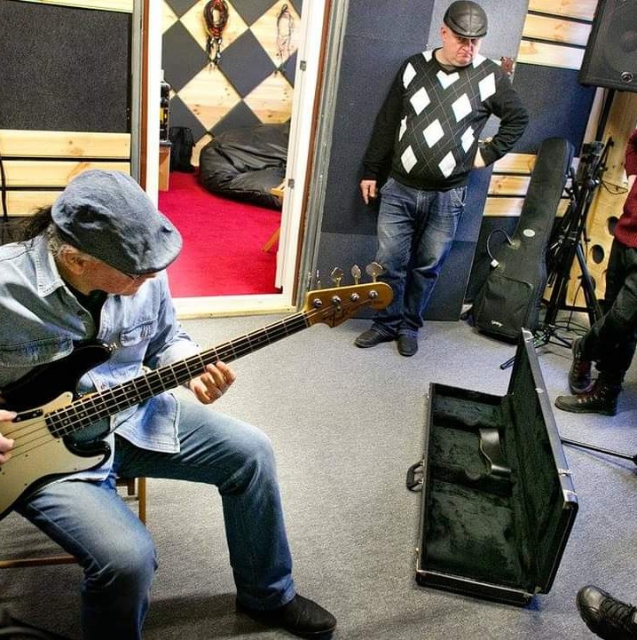 Muzyk grający na gitarze basowej w studio nagraniowym z czerwonym dywanem w tle, obok otwarty futerał na instrument i stojący mężczyzna w swetrze w romby.