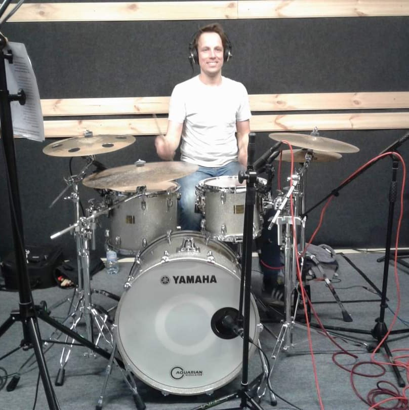 Uśmiechnięty perkusista w słuchawkach siedzi za zestawem perkusyjnym Yamaha w szarym studio, trzymając pałeczki w dłoniach.