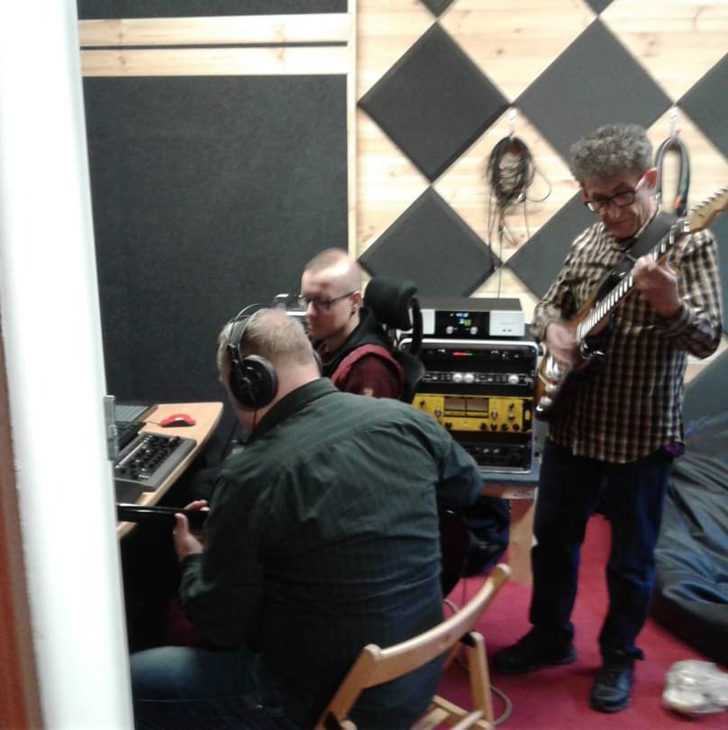 Trzech mężczyzn w studiu nagraniowym; jeden gra na gitarze elektrycznej, drugi siedzi przy mikserze w słuchawkach, trzeci siedzi w tle; ściany wyłożone panelami akustycznymi.