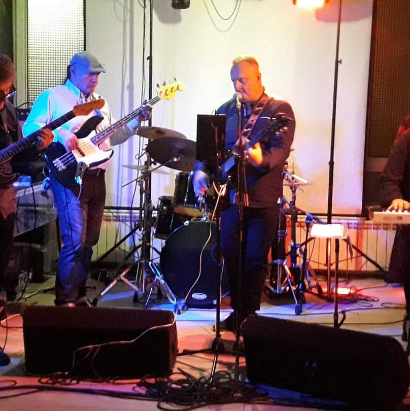 Zespół muzyczny w trakcie występu: gitarzysta basowy w czapce, wokalista z gitarą, perkusja w tle, kable na podłodze.