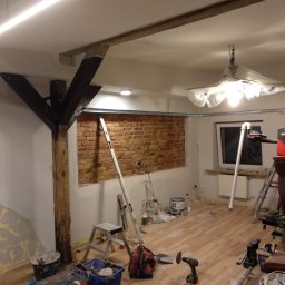 Uslugi ogólnobudowlane Jagielnicki Jaroslaw - Pokój w trakcie prac wykończeniowych: odsłonięta ceglana ściana, drewniane belki stropowe, podłoga z paneli, narzędzia i materiały budowlane rozłożone na podłodze, lampa zabezpieczona folią.