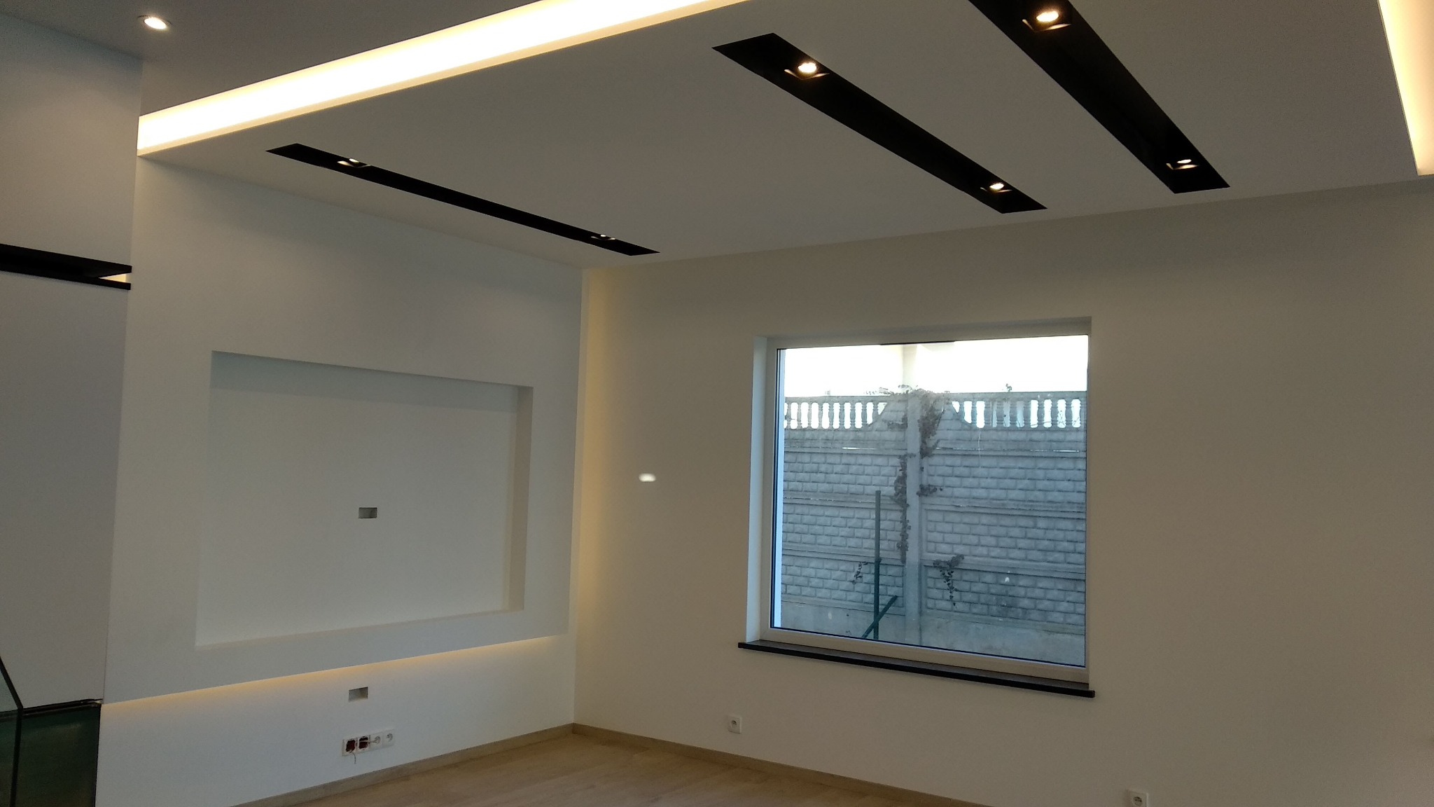 Nowoczesne wnętrze z białymi ścianami, drewnianą podłogą i sufitem podwieszanym z oświetleniem LED oraz czarnymi listwami z reflektorami punktowymi, w tle okno z widokiem na betonowy płot.