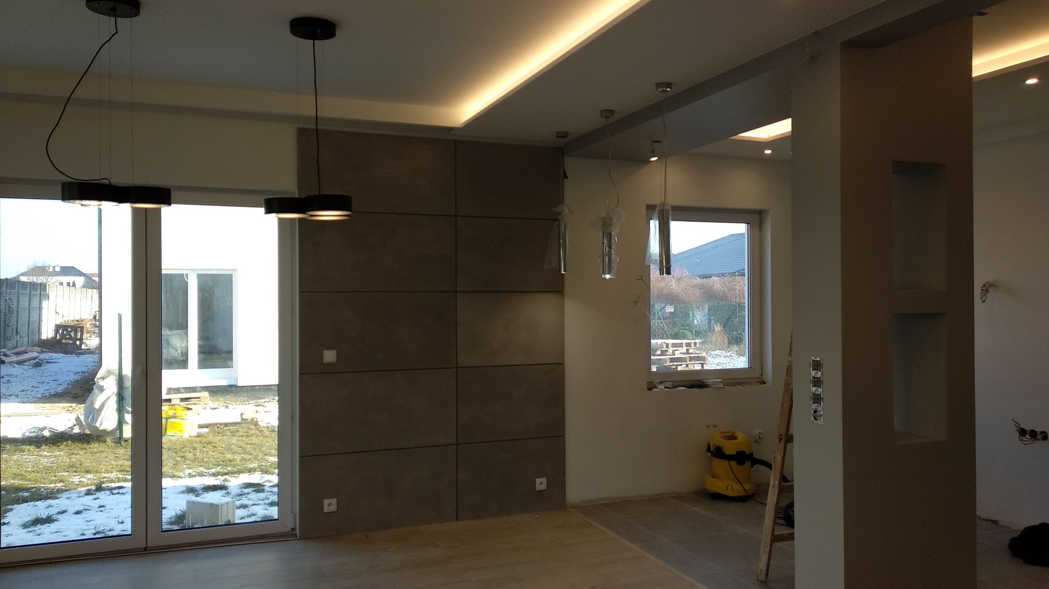 Nowoczesne wnętrze w trakcie wykańczania: panelowa ściana z szarego betonu, sufit z oświetleniem LED, podwieszane lampy z czarnymi kloszami, widoczne okno z widokiem na ośnieżony ogród, odkurzacz...