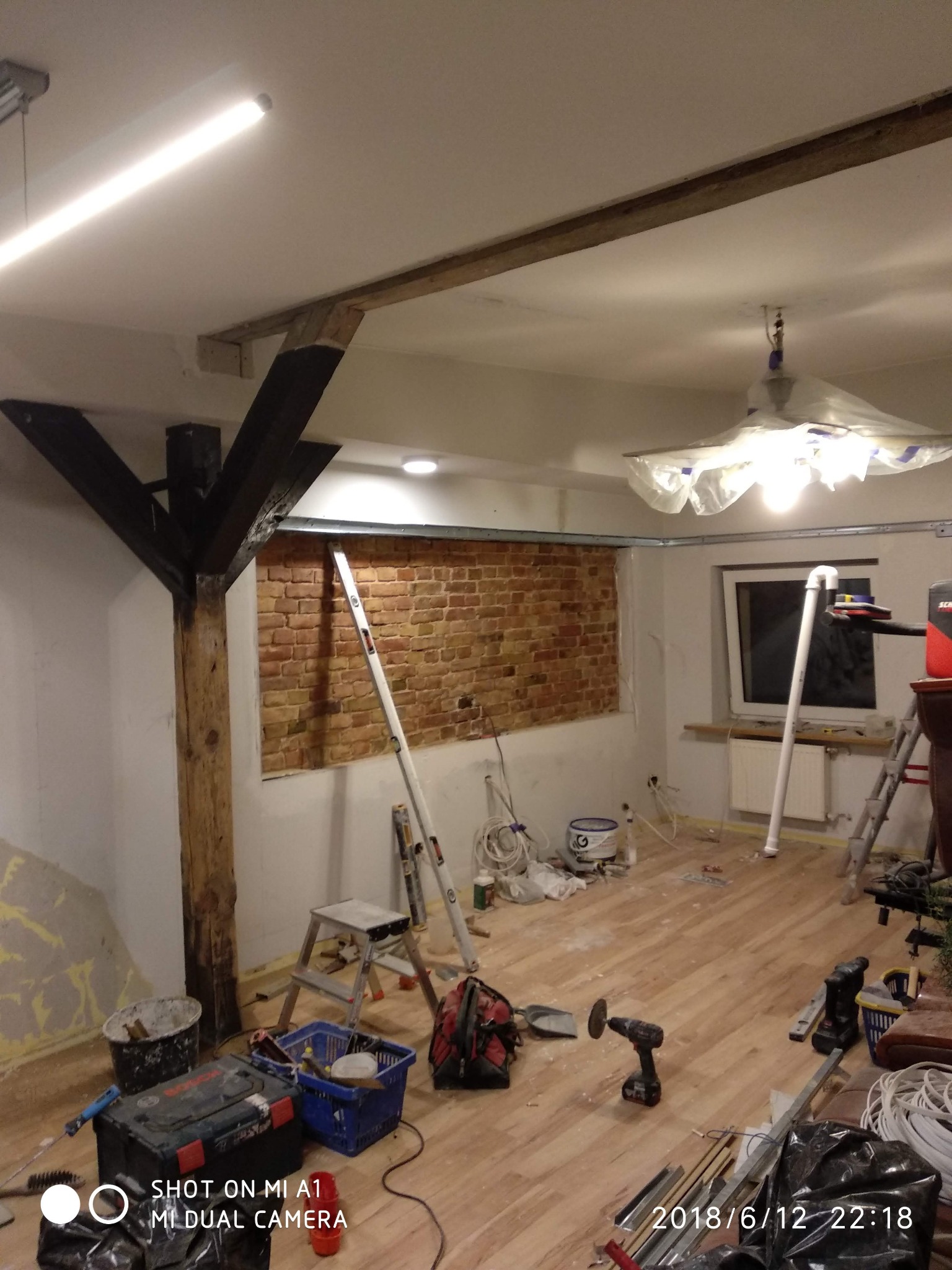 Pokój w trakcie prac wykończeniowych: odsłonięta ceglana ściana, drewniane belki stropowe, podłoga z paneli, narzędzia i materiały budowlane rozłożone na podłodze, lampa zabezpieczona folią.