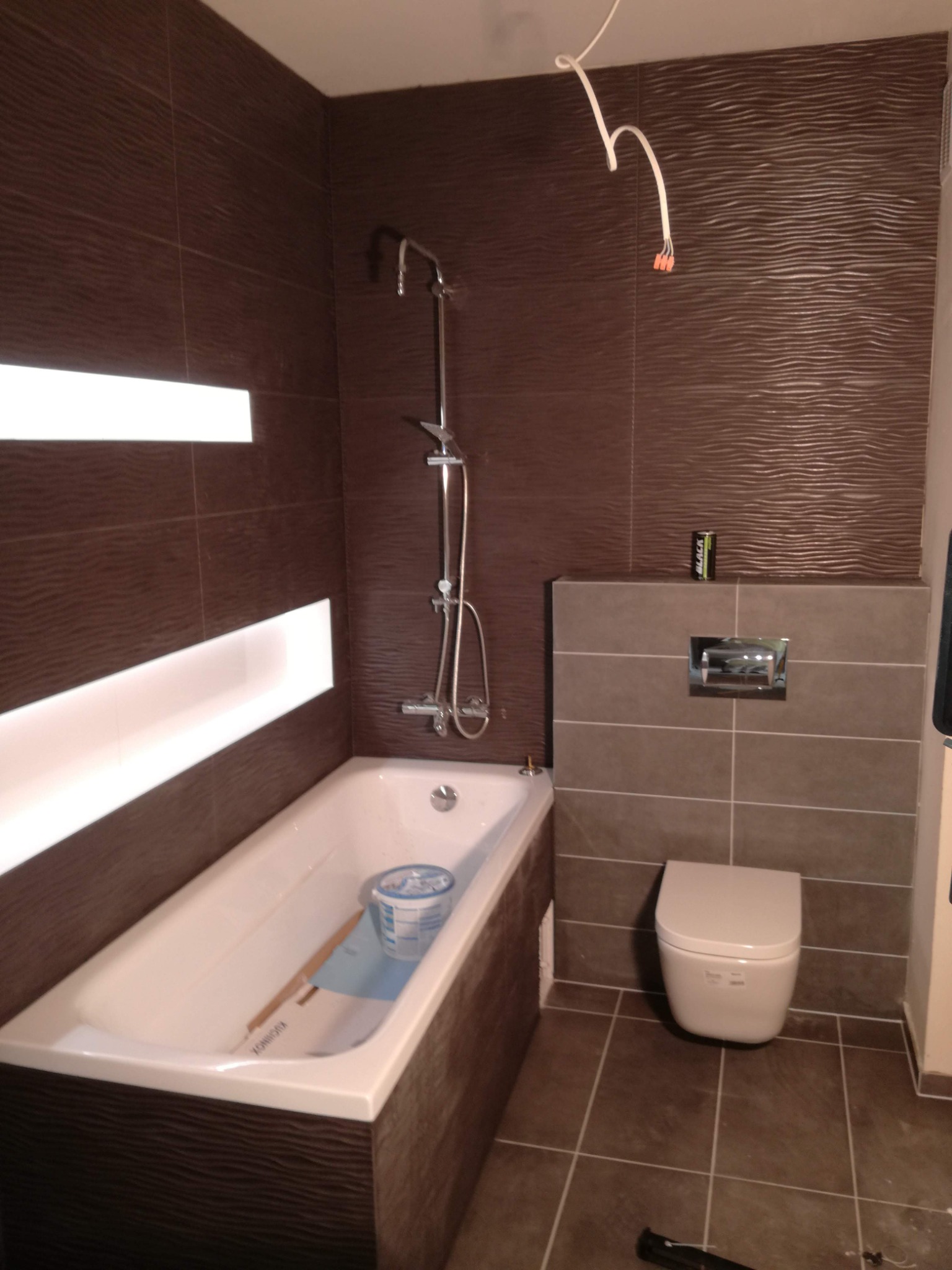Wnętrze łazienki w trakcie prac wykończeniowych, widoczna wanna z zabezpieczeniem i pojemnikiem, toaleta, brązowe płytki z falistą strukturą na ścianach, biała listwa LED, wystające przewody...