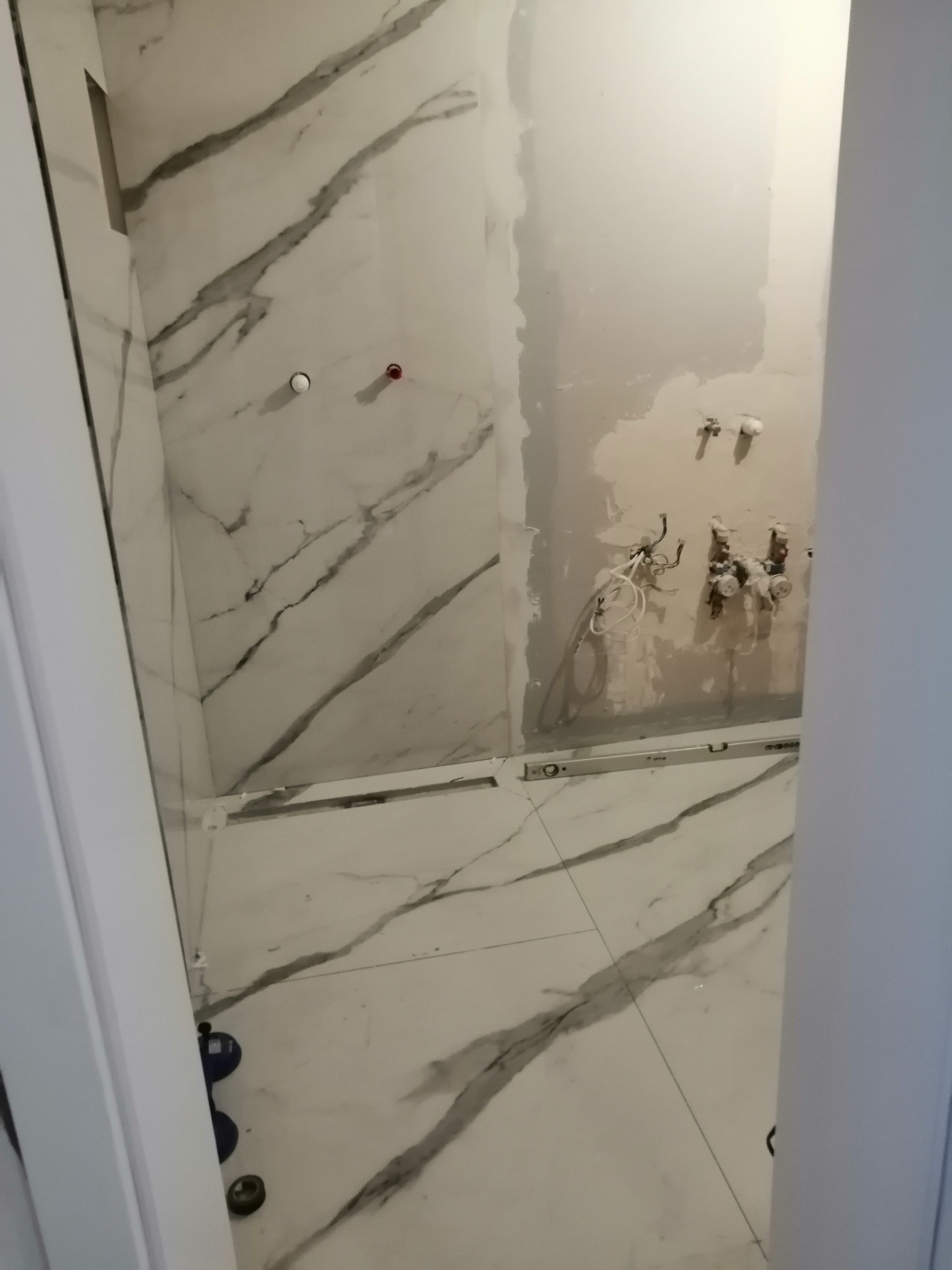 Wnętrze łazienki w trakcie prac wykończeniowych, widoczne białe płytki z szarym wzorem żyłowania na ścianach i podłodze, przygotowane instalacje wodne i poziomica. Część ściany jest w stanie...