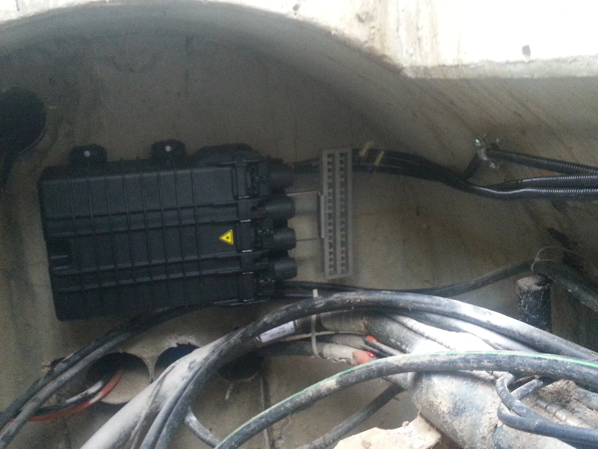 Czarna skrzynka telekomunikacyjna z żółtym trójkątem ostrzegawczym, podłączona do kabli w betonowej studzience kanalizacyjnej.