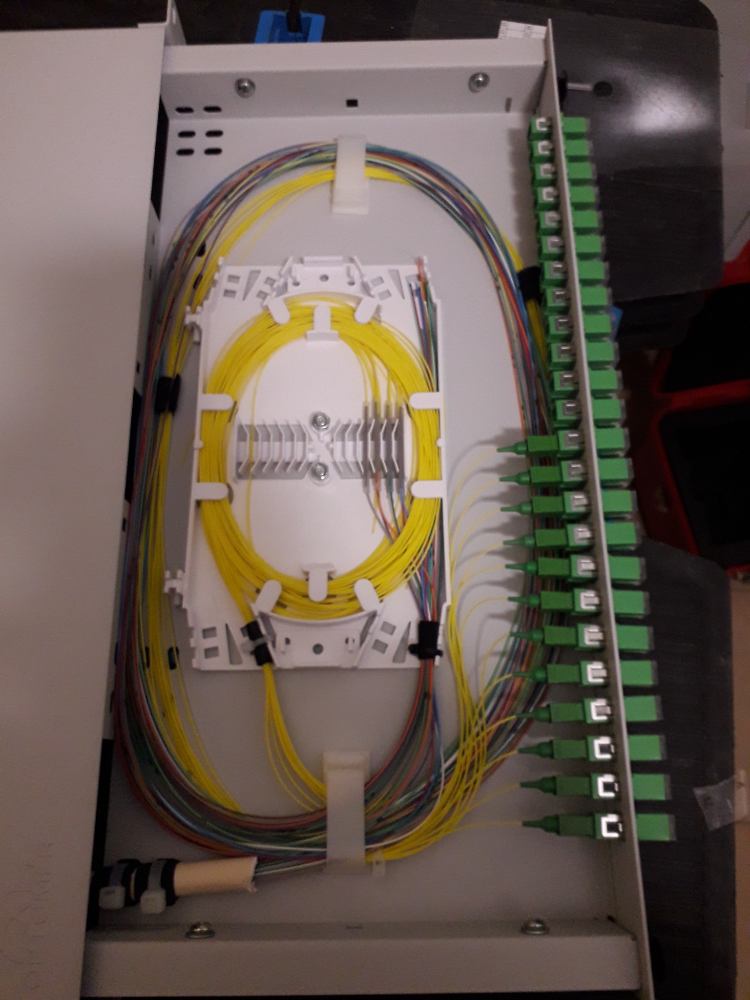 Panel światłowodowy z uporządkowanymi kablami w różnych kolorach, zakończonymi zielonymi złączkami, umieszczony w metalowej obudowie.