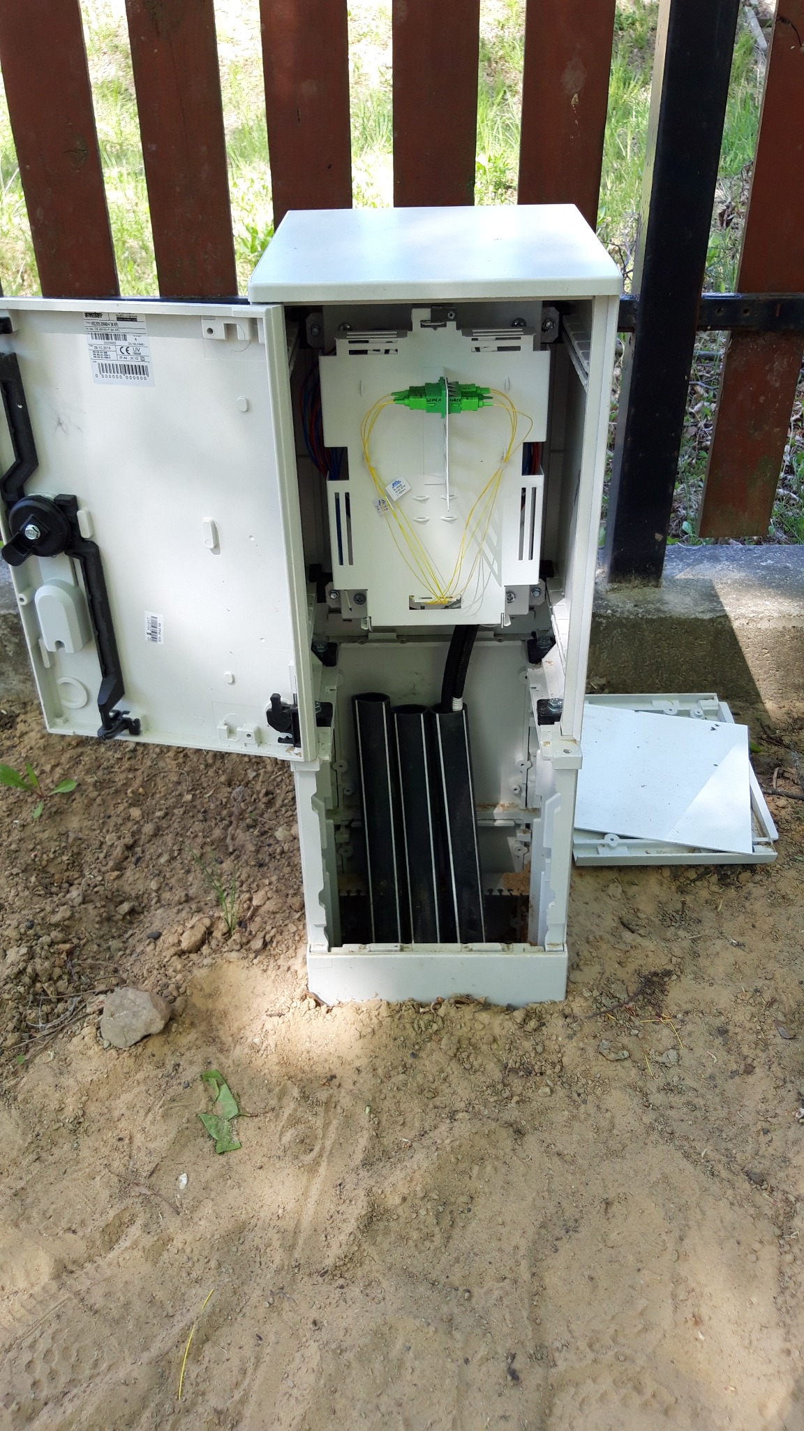 Otwarta skrzynka telekomunikacyjna z widocznymi kablami światłowodowymi i elementami łączeniowymi, umieszczona na ziemi, obok drewnianego ogrodzenia.