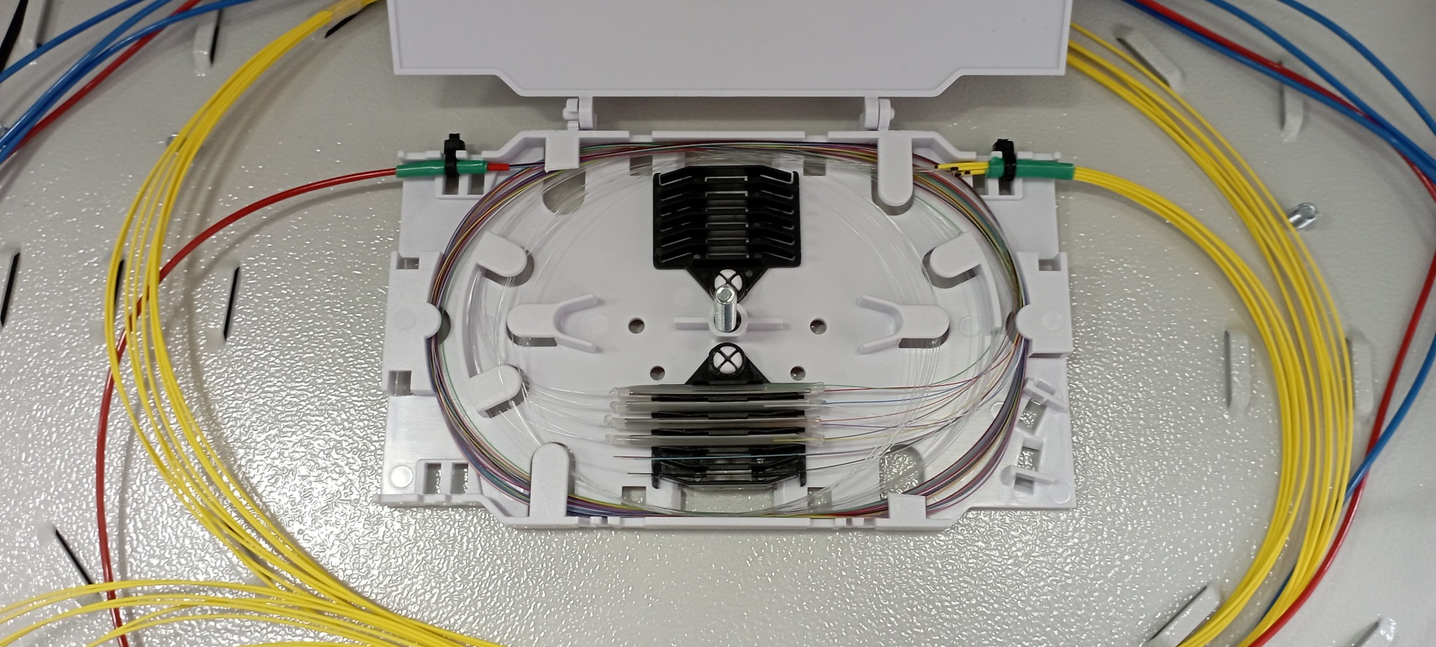 Otwarta, biała skrzynka światłowodowa z widocznymi, uporządkowanymi kablami światłowodowymi w różnych kolorach, ułożonymi w pętle wewnątrz obudowy.