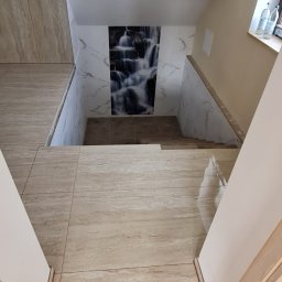 Usługi remontowo budowlane - Klatka schodowa w Skawie wyłożona beżowymi płytkami imitującymi trawertyn, z dekoracyjnym panelem z motywem wodospadu na ścianie. Widoczne niedokończone prace przy podłodze.