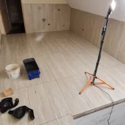 Podłoga i ściany w poddaszu wyłożone beżowymi płytkami imitującymi marmur, widoczny halogen na statywie, wiadro i kuweta malarska oraz nakolanniki, etap prac wykończeniowych.