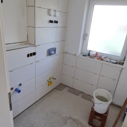 Usługi remontowo budowlane - Białe płytki ceramiczne na ścianach łazienki w trakcie montażu, widoczne krzyżyki dystansowe, przygotowane otwory na armaturę, surowy beton na podłodze, wiadro z wodą na plastikowej skrzynce, okno...