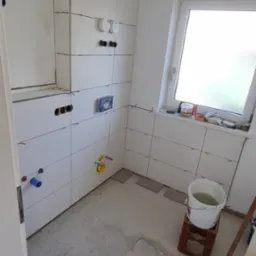 Białe płytki ceramiczne na ścianach łazienki w trakcie montażu, widoczne krzyżyki dystansowe, przygotowane otwory na armaturę, surowy beton na podłodze, wiadro z wodą na plastikowej skrzynce, okno...