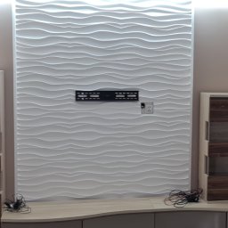 Usługi remontowo budowlane - Nowoczesna ściana z paneli 3D w odcieniach bieli, z zamontowanym uchwytem na telewizor i gniazdkiem elektrycznym, nad niską szafką RTV z zaokrąglonymi frontami, uzupełniona o dekoracyjny żyrandol.