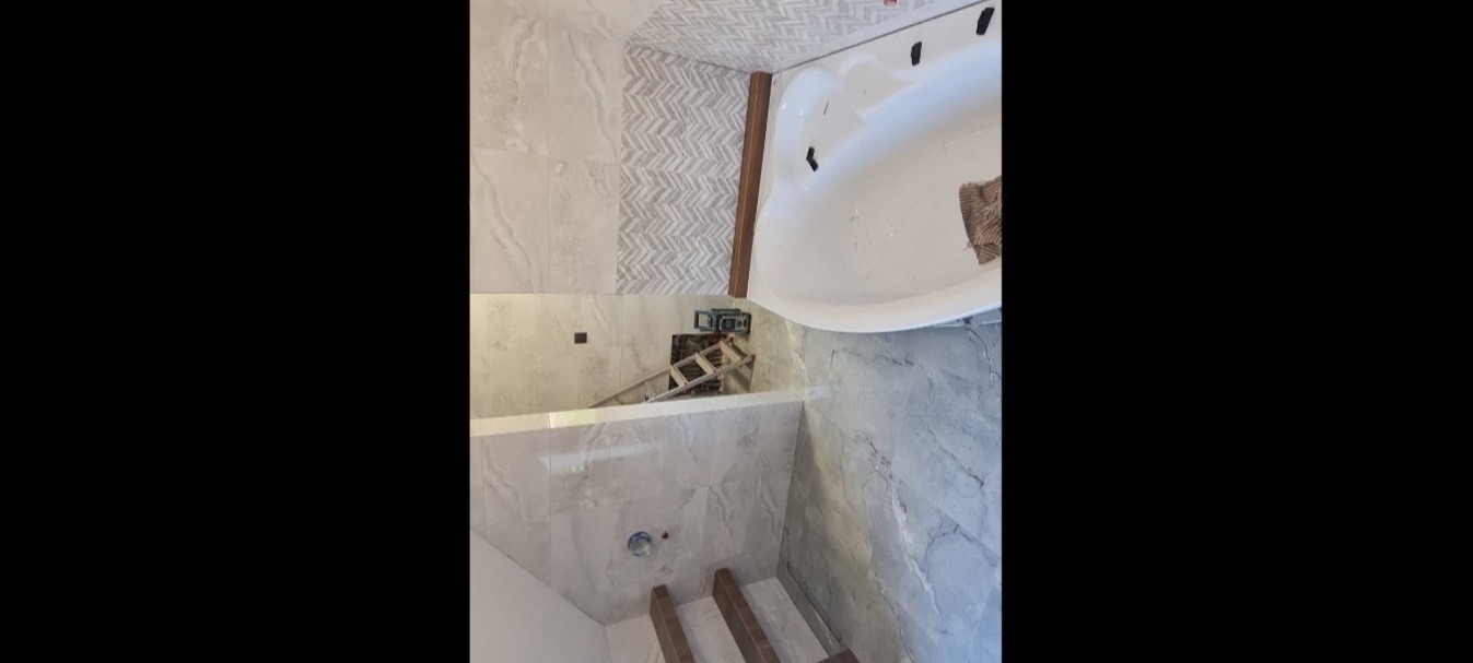 Fragment łazienki w trakcie remontu, widoczna wanna narożna, płytki imitujące marmur na ścianach, drabina rozstawiona przy ścianie, przygotowane podłoże pod podłogę.