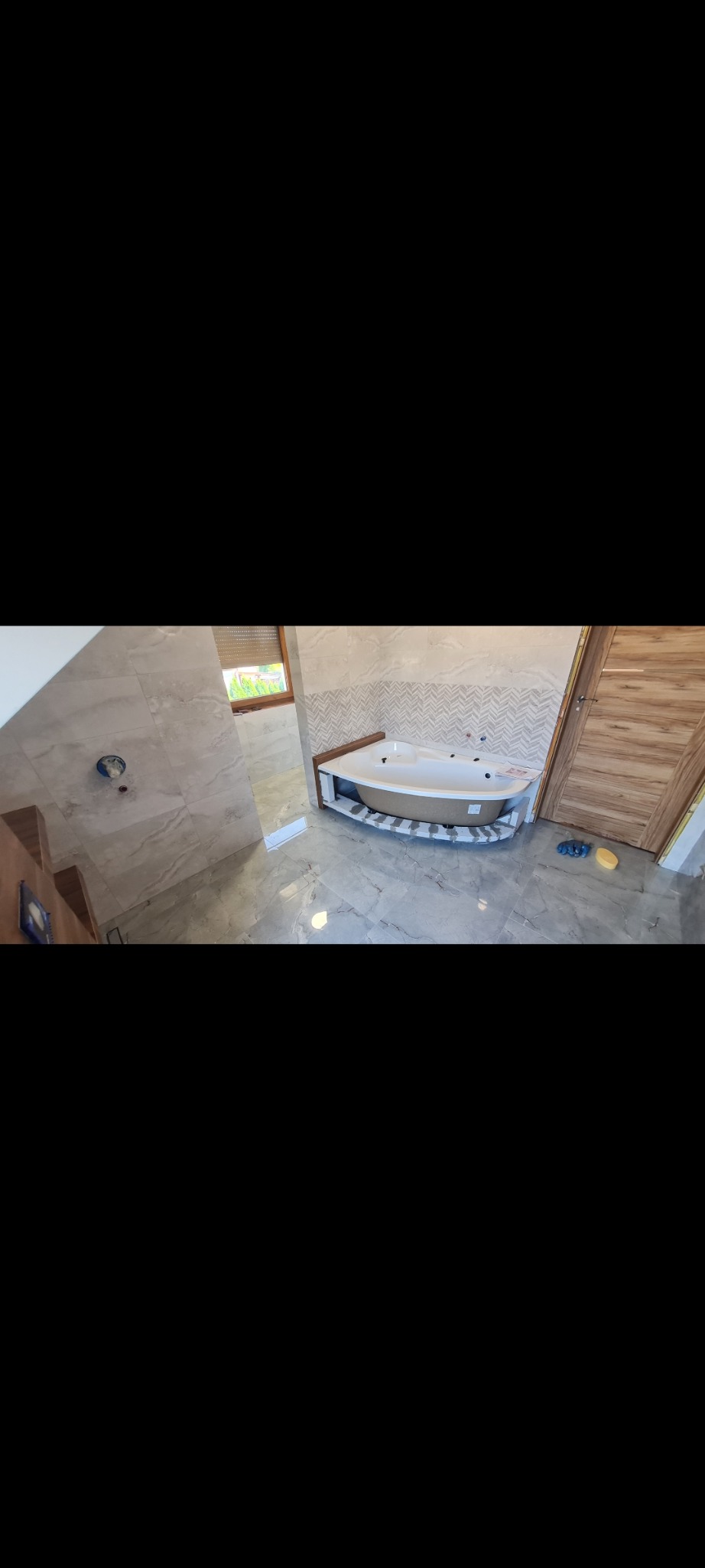 Narożna wanna z hydromasażem w trakcie montażu, obudowana płytkami imitującymi drewno, na podłodze i ścianach szare płytki marmurowe, widoczne narzędzia i materiały budowlane.