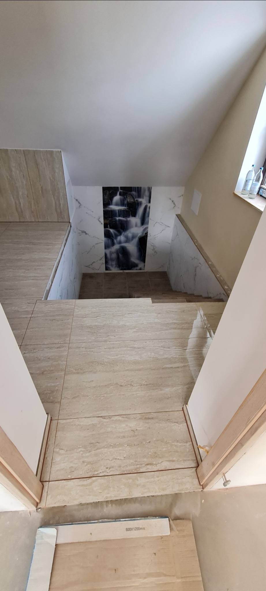 Klatka schodowa w Skawie wyłożona beżowymi płytkami imitującymi trawertyn, z dekoracyjnym panelem z motywem wodospadu na ścianie. Widoczne niedokończone prace przy podłodze.