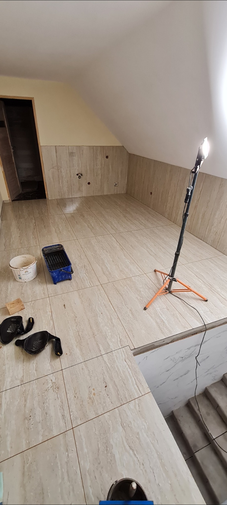 Podłoga i ściany w poddaszu wyłożone beżowymi płytkami imitującymi marmur, widoczny halogen na statywie, wiadro i kuweta malarska oraz nakolanniki, etap prac wykończeniowych.