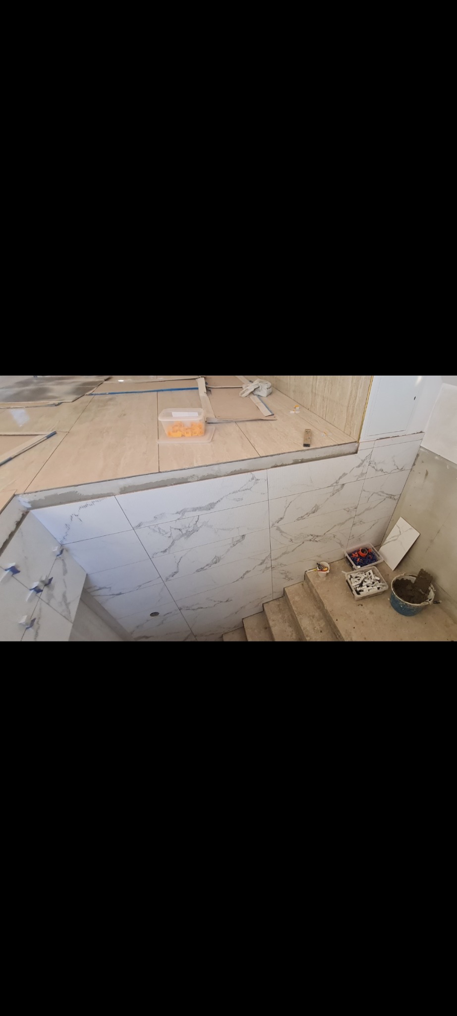 Układanie płytek imitujących marmur na ścianach i podłodze w łazience, widoczne narzędzia i materiały budowlane na schodach.