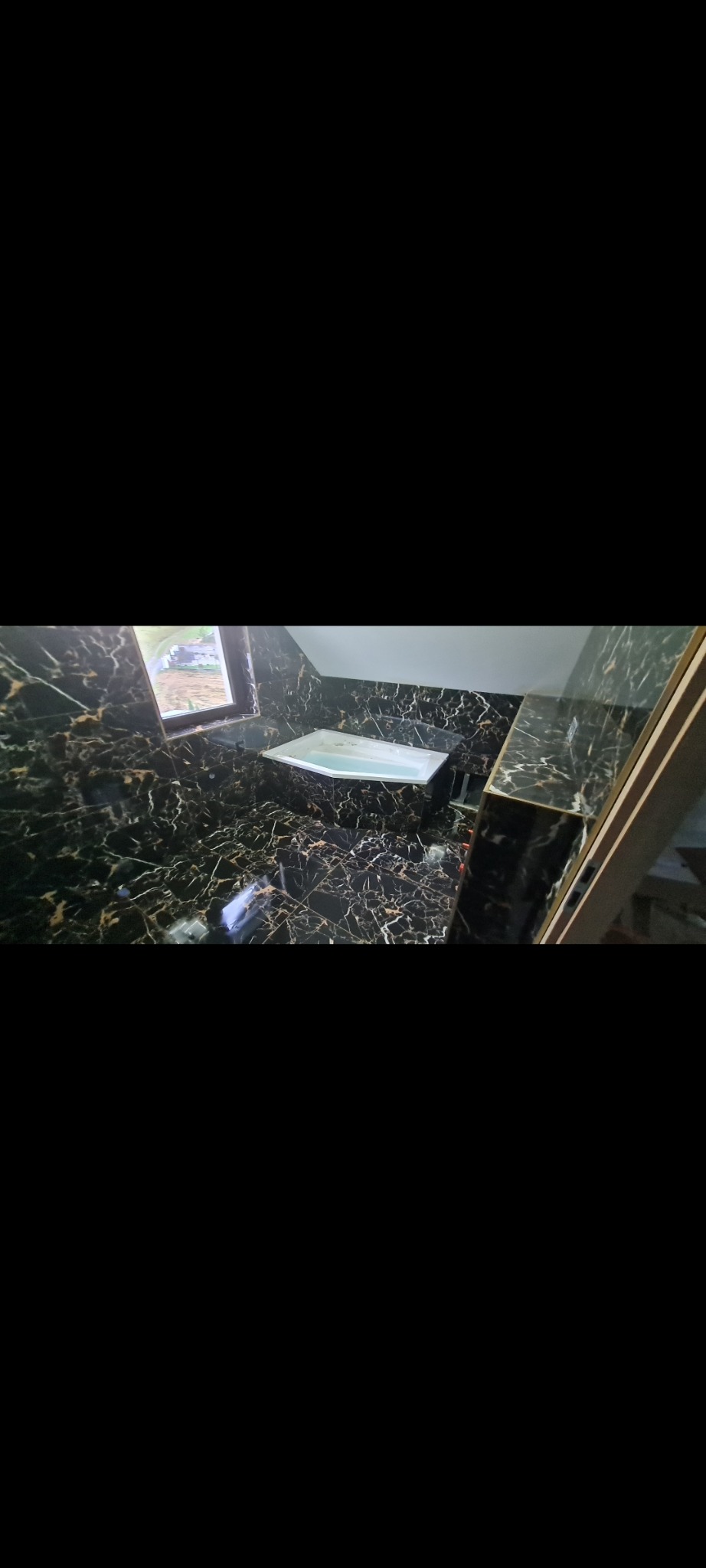 Błyszcząca, czarna łazienka z marmurowym wzorem, z białą umywalką pod oknem i częściowo wykończoną ścianą po prawej stronie.