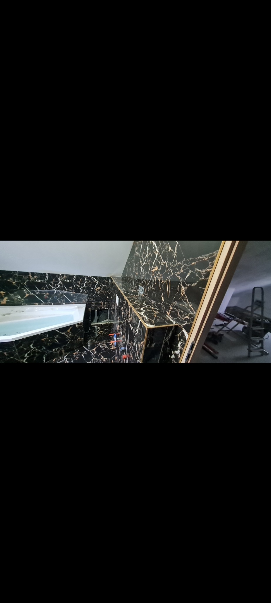 Nowoczesna łazienka z czarnymi, marmurkowymi płytkami, złotymi akcentami i białą wanną, widok na fragment pomieszczenia z drabinką i narzędziami w tle.