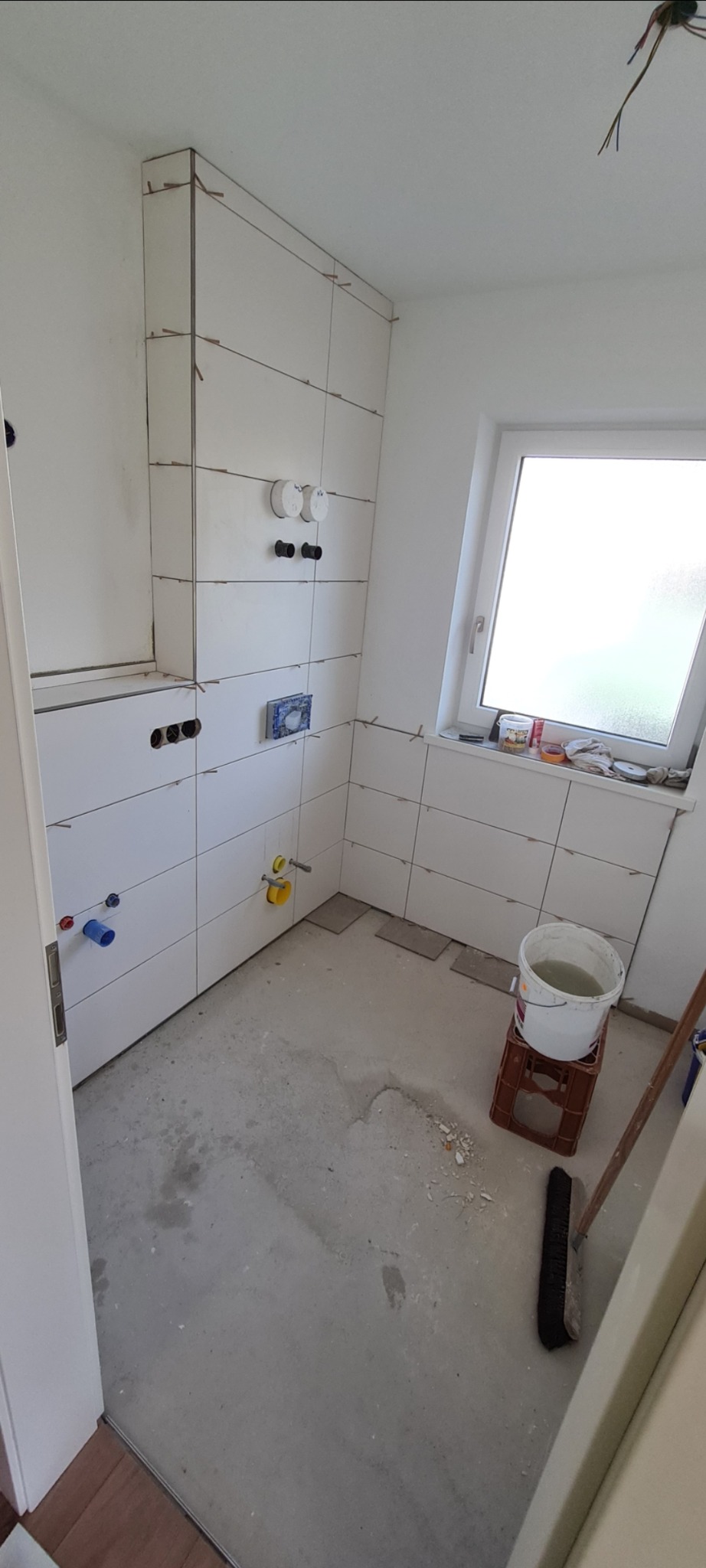 Białe płytki ceramiczne na ścianach łazienki w trakcie montażu, widoczne krzyżyki dystansowe, przygotowane otwory na armaturę, surowy beton na podłodze, wiadro z wodą na plastikowej skrzynce, okno...