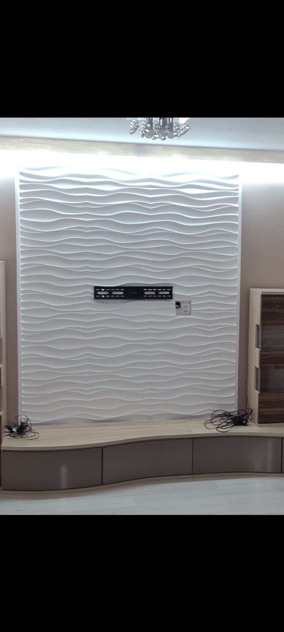 Nowoczesna ściana z paneli 3D w odcieniach bieli, z zamontowanym uchwytem na telewizor i gniazdkiem elektrycznym, nad niską szafką RTV z zaokrąglonymi frontami, uzupełniona o dekoracyjny żyrandol.