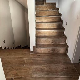 Sebastian Buczak usługi ogólnobudowlane - Wykończone schody wewnętrzne wyłożone brązowymi płytkami imitującymi drewno, z widocznym fragmentem podłogi również wyłożonej tymi samymi płytkami, ściany pomalowane na biało z zainstalowanymi...