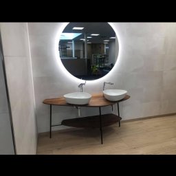 Sebastian Buczak usługi ogólnobudowlane - Nowoczesna łazienka z podwójną umywalką w kształcie misy na drewnianym blacie z metalowymi nogami, okrągłe lustro z podświetleniem LED, jasne płytki na ścianach i drewnopodobna podłoga.