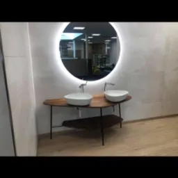 Nowoczesna łazienka z podwójną umywalką w kształcie misy na drewnianym blacie z metalowymi nogami, okrągłe lustro z podświetleniem LED, jasne płytki na ścianach i drewnopodobna podłoga.