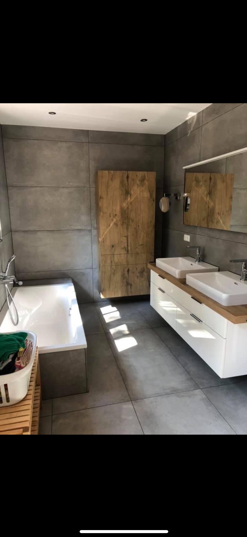 Nowoczesna łazienka z szarymi płytkami wielkoformatowymi, drewnianą szafką, białą podwójną umywalką na drewnianym blacie i wanną z przymocowanym prysznicem, oświetlona promieniami słońca.