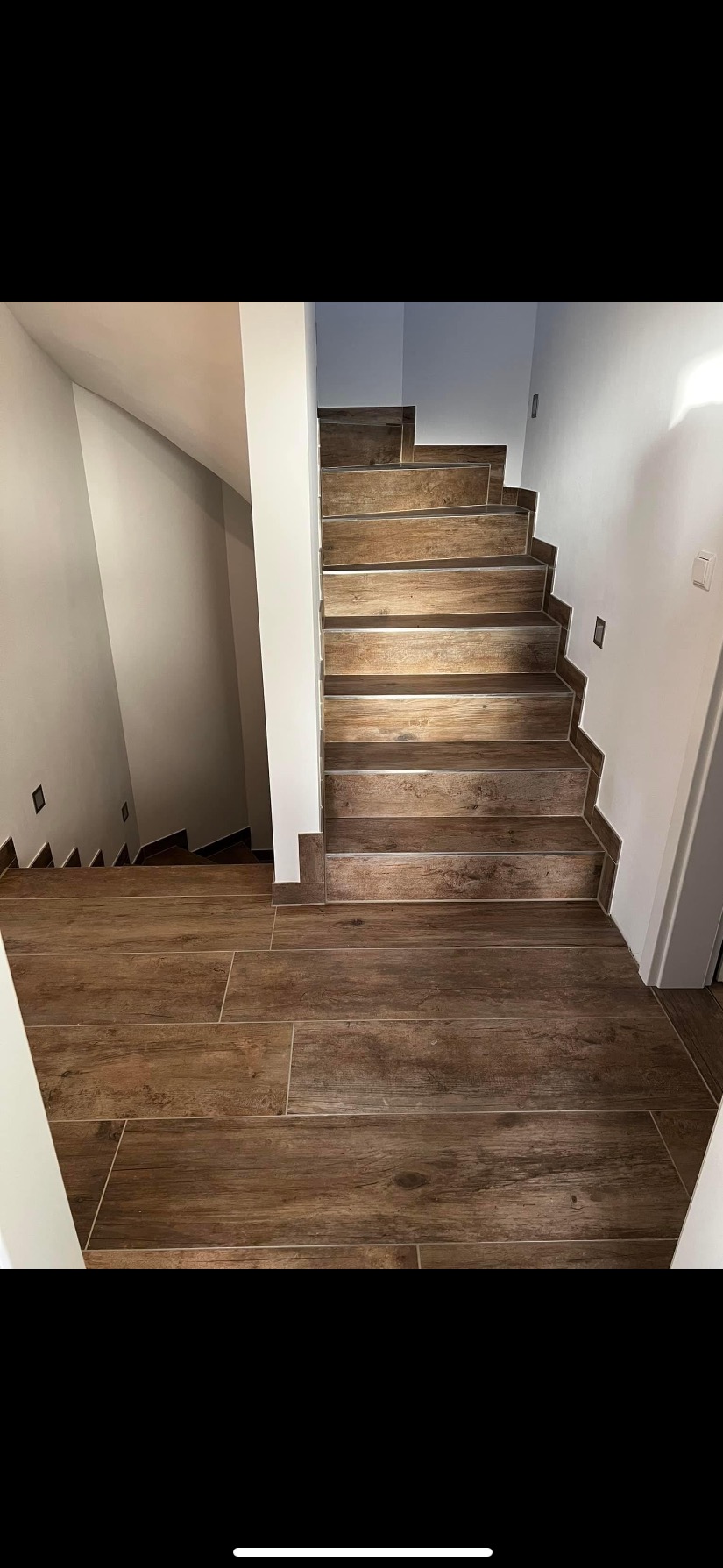 Wykończone schody wewnętrzne wyłożone brązowymi płytkami imitującymi drewno, z widocznym fragmentem podłogi również wyłożonej tymi samymi płytkami, ściany pomalowane na biało z zainstalowanymi...