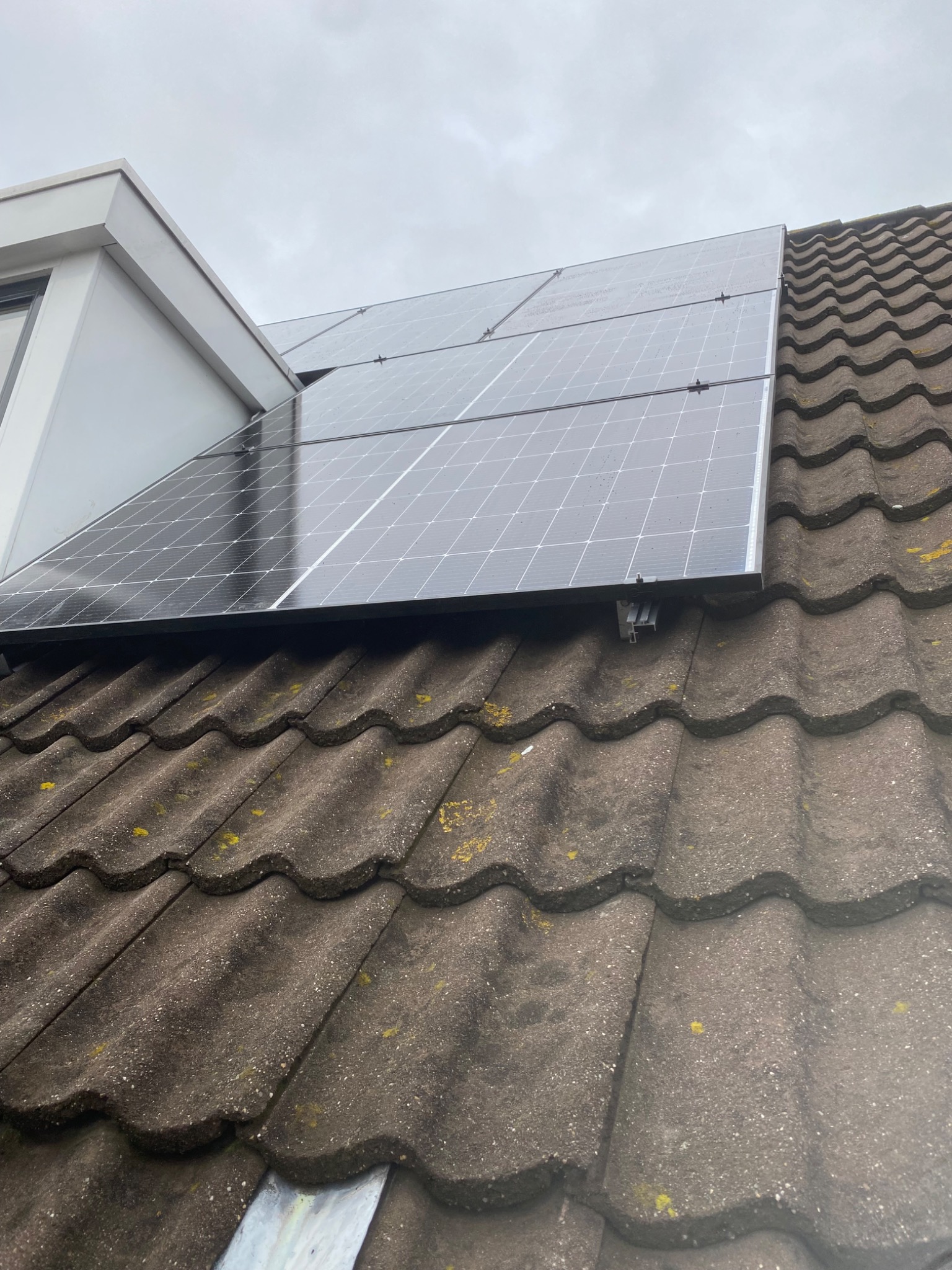 Panele słoneczne zamontowane na dachu z ciemnej dachówki ceramicznej, widoczne mocowania i fragment okna dachowego przy pochmurnym niebie.
