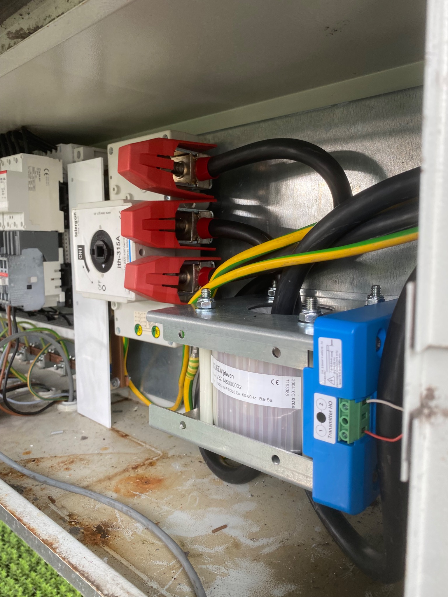 Wnętrze skrzynki elektrycznej z widocznymi przewodami, bezpiecznikami z czerwonymi osłonami, transformatorem i niebieskim modułem nadajnika, zardzewiałe elementy metalowe.