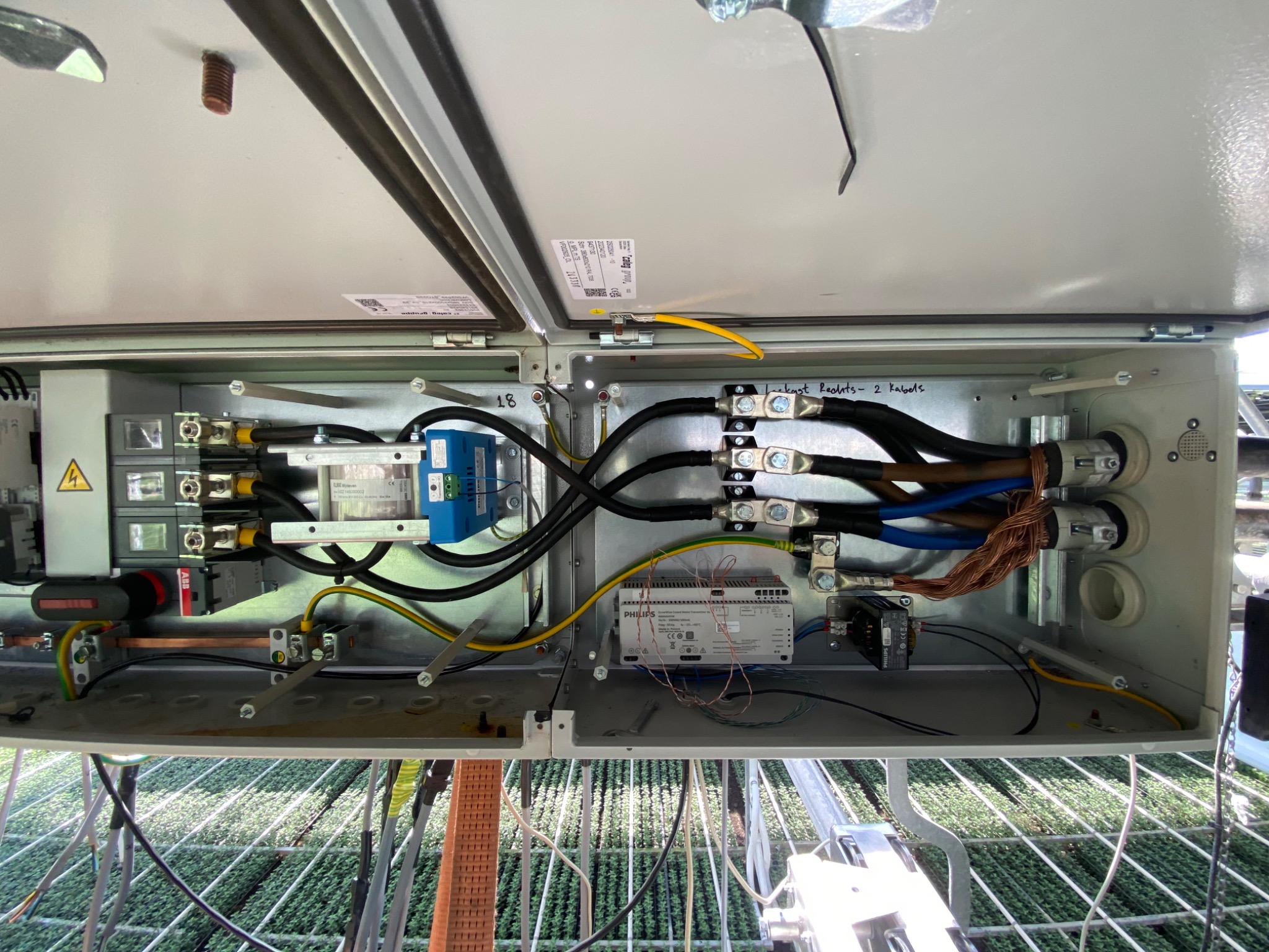 Otwarta skrzynka elektryczna z widocznymi bezpiecznikami, przewodami o różnych kolorach, transformatorem i modułem Philips, zamontowana wewnątrz konstrukcji szklarni.