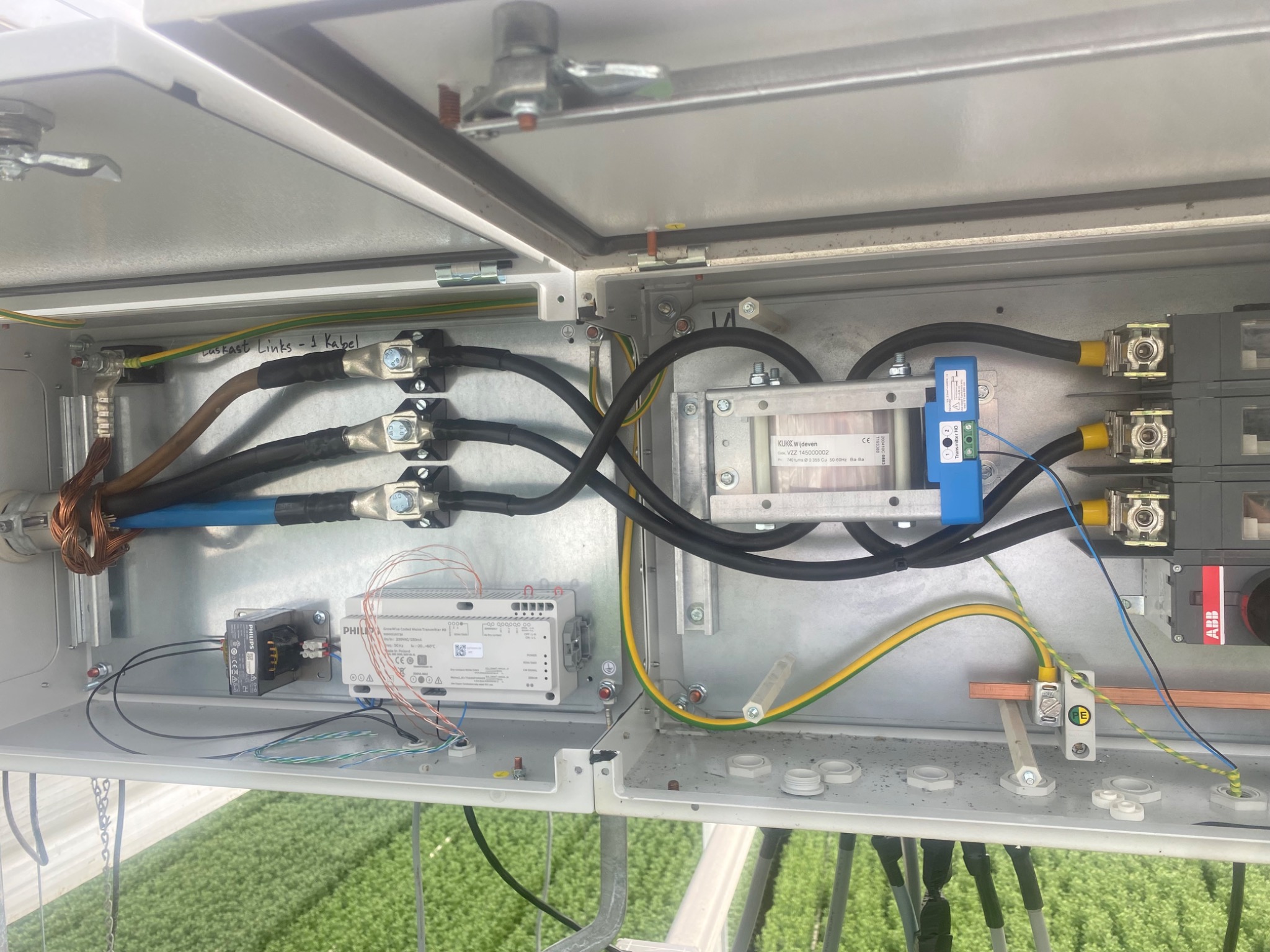 Otwarta skrzynka elektryczna z widocznymi przewodami, złączkami, sterownikami Philips i elementami ABB, zainstalowana na zewnątrz z zielenią w tle.