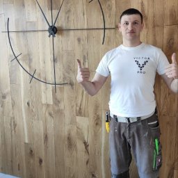 ViktorPro - Mężczyzna w roboczym ubraniu prezentuje zamontowany na drewnianej ścianie minimalistyczny, czarny zegar bez cyfr, dając kciuki w górę.