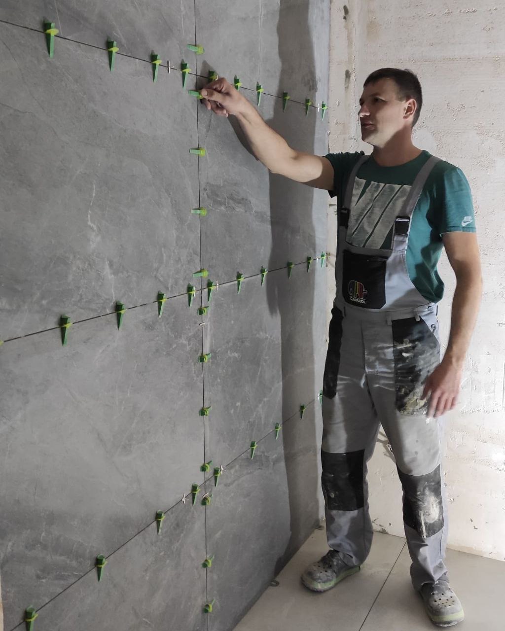 Mężczyzna w kombinezonie roboczym, z logo firmy Caparol, układa płytki imitujące marmur, używając systemu poziomowania z zielonymi klinami, na ścianie w surowym stanie.