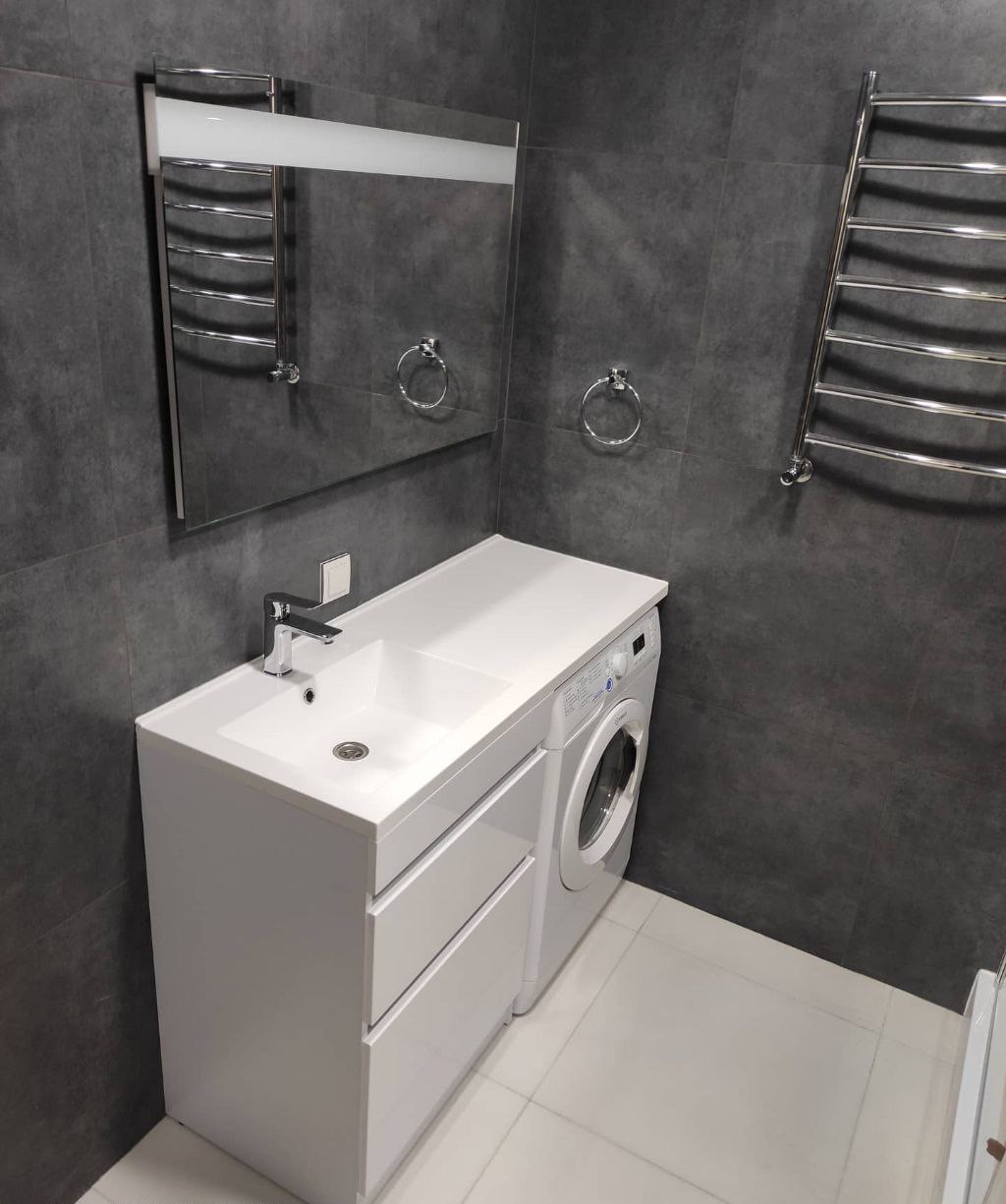 Nowoczesna łazienka z szarymi płytkami, białą umywalką z szafką, pralką w zabudowie, podświetlanym lustrem i grzejnikiem drabinkowym.