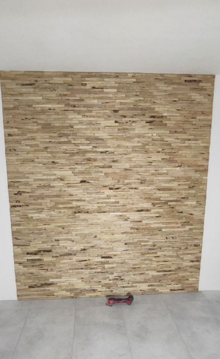 Ściana wykończona panelami z naturalnego drewna o nieregularnej strukturze, ułożonymi poziomo, z widocznym narzędziem wielofunkcyjnym na podłodze.