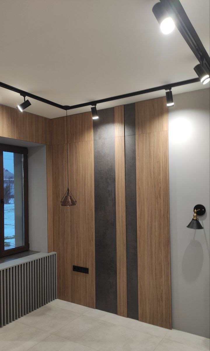 Nowoczesne wnętrze z panelami ściennymi w odcieniach drewna i szarości, oświetlenie szynowe z reflektorami punktowymi i wisząca lampa druciana, grzejnik dekoracyjny z pionowymi lamelami.