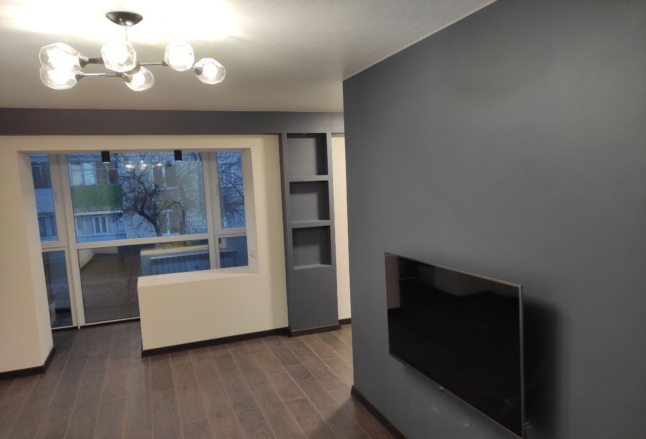 Nowoczesny salon po remoncie: szara ściana z telewizorem, drewniana podłoga, okno z widokiem na budynek, designerska lampa sufitowa.