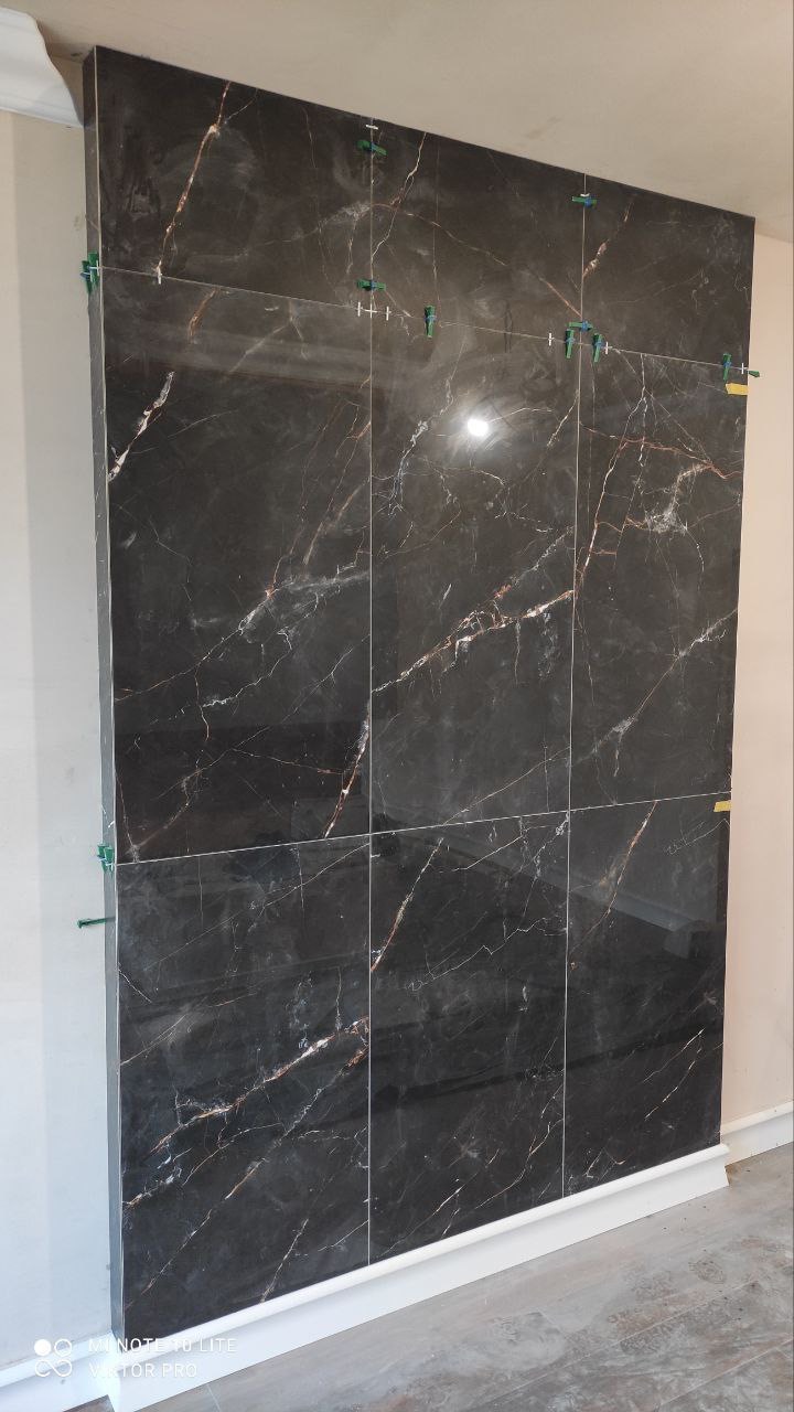 Montaż płytek imitujących czarny marmur na ścianie, widoczne plastikowe krzyżyki dystansowe, jasne fugi, biała listwa przypodłogowa.