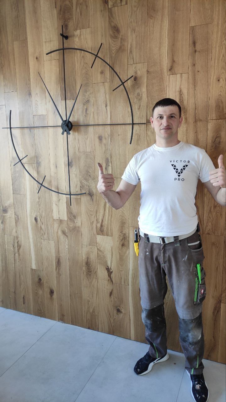 Mężczyzna w roboczym ubraniu prezentuje zamontowany na drewnianej ścianie, minimalistyczny zegar ścienny z metalowymi wskazówkami i oznaczeniami godzin, unosząc kciuki w górę.