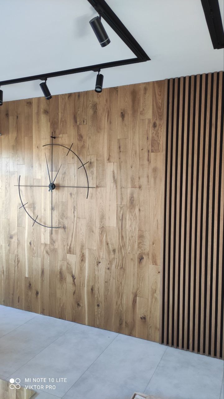 Drewniana ściana z panelami pionowymi i zegarem w stylu minimalistycznym, oświetlona czarnymi reflektorami sufitowymi.