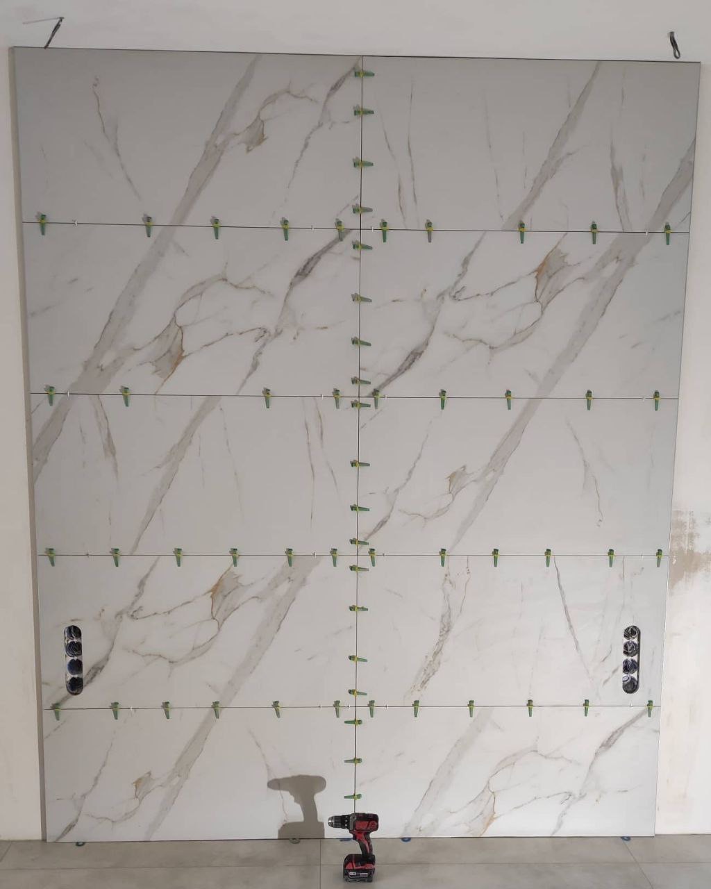 Montaż wielkoformatowych płytek imitujących marmur na ścianie, z widocznymi zielonymi krzyżykami poziomującymi i puszkami elektrycznymi, w tle wiertarko-wkrętarka.