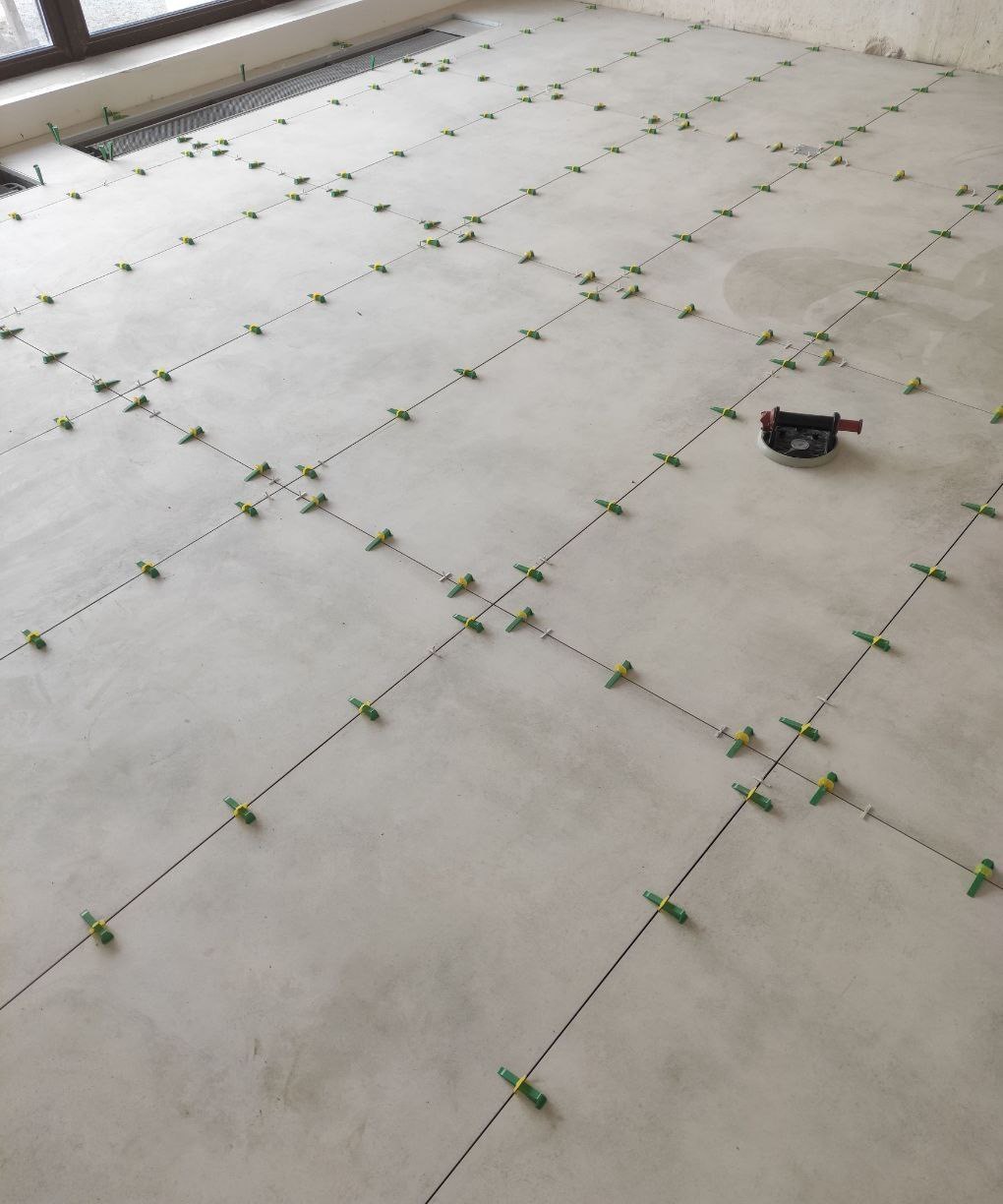 Przygotowanie do układania płytek: wyznaczona siatka z krzyżykami poziomującymi na szarej wylewce, widoczna przyssawka do przenoszenia płytek.